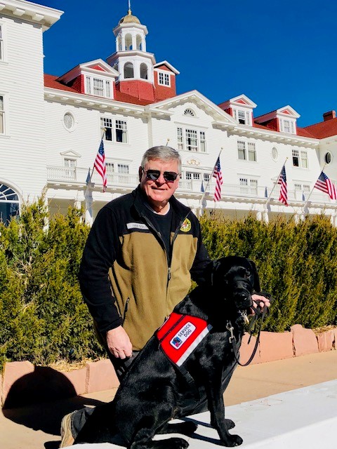 man and service dog in red vest in front of large white building
